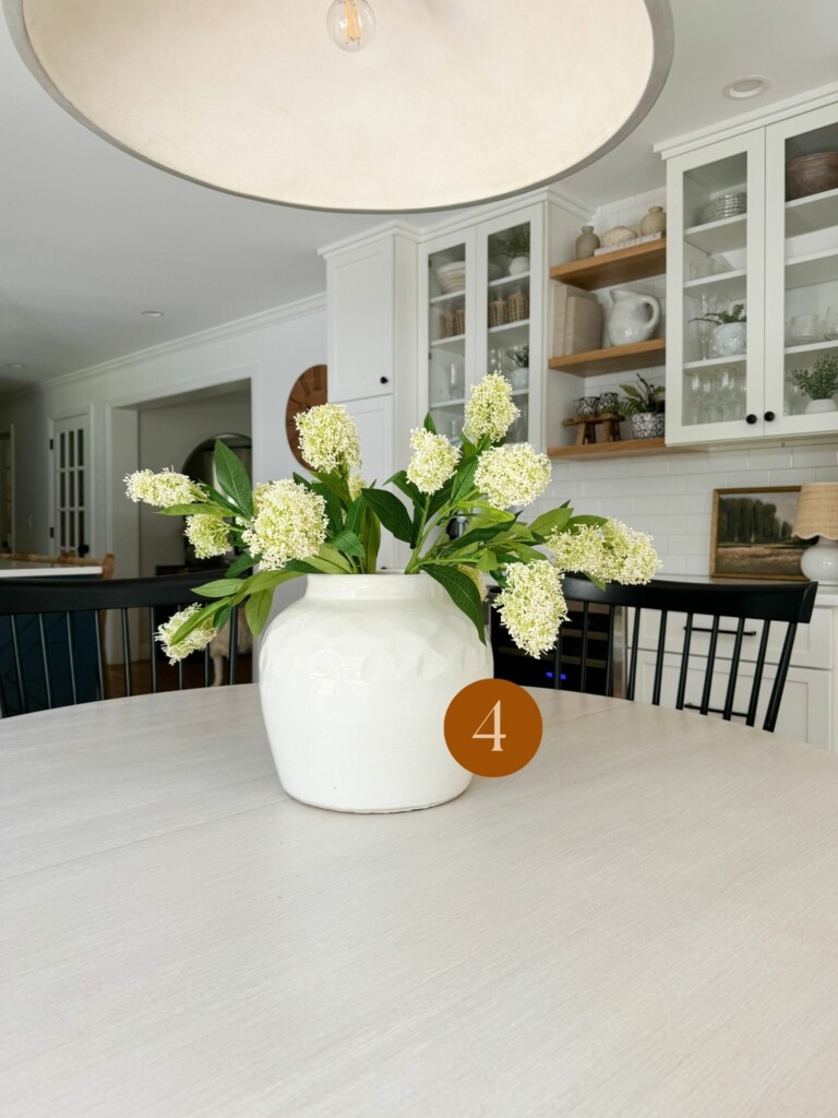 Bright coastal kitchen with a round dining table styled with a white ceramic vase and fresh green floral centerpiece.