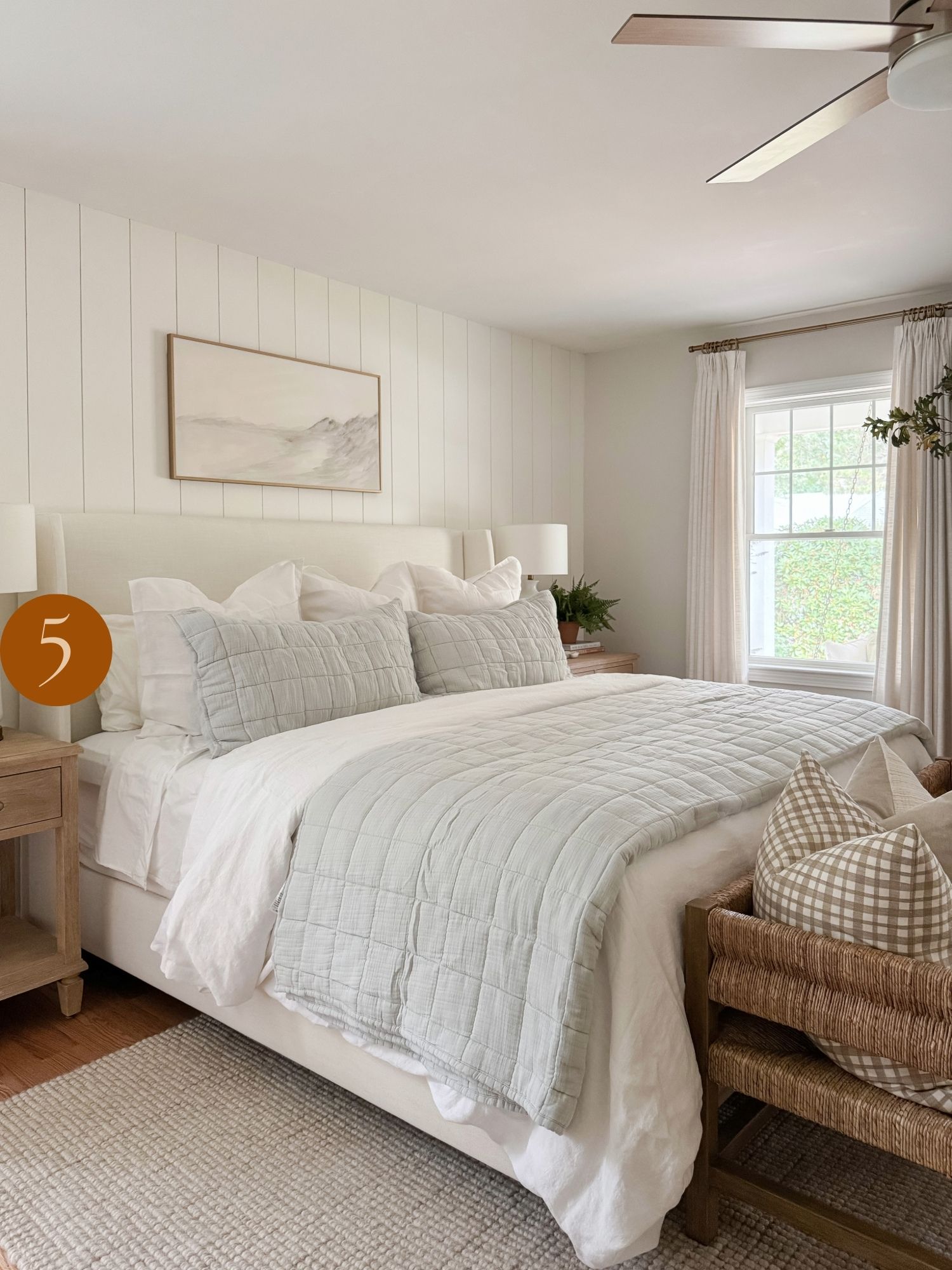 neutral living room styling inspiration Relaxing coastal bedroom with white shiplap accent wall, upholstered bed, light blue quilt, and neutral layered bedding.