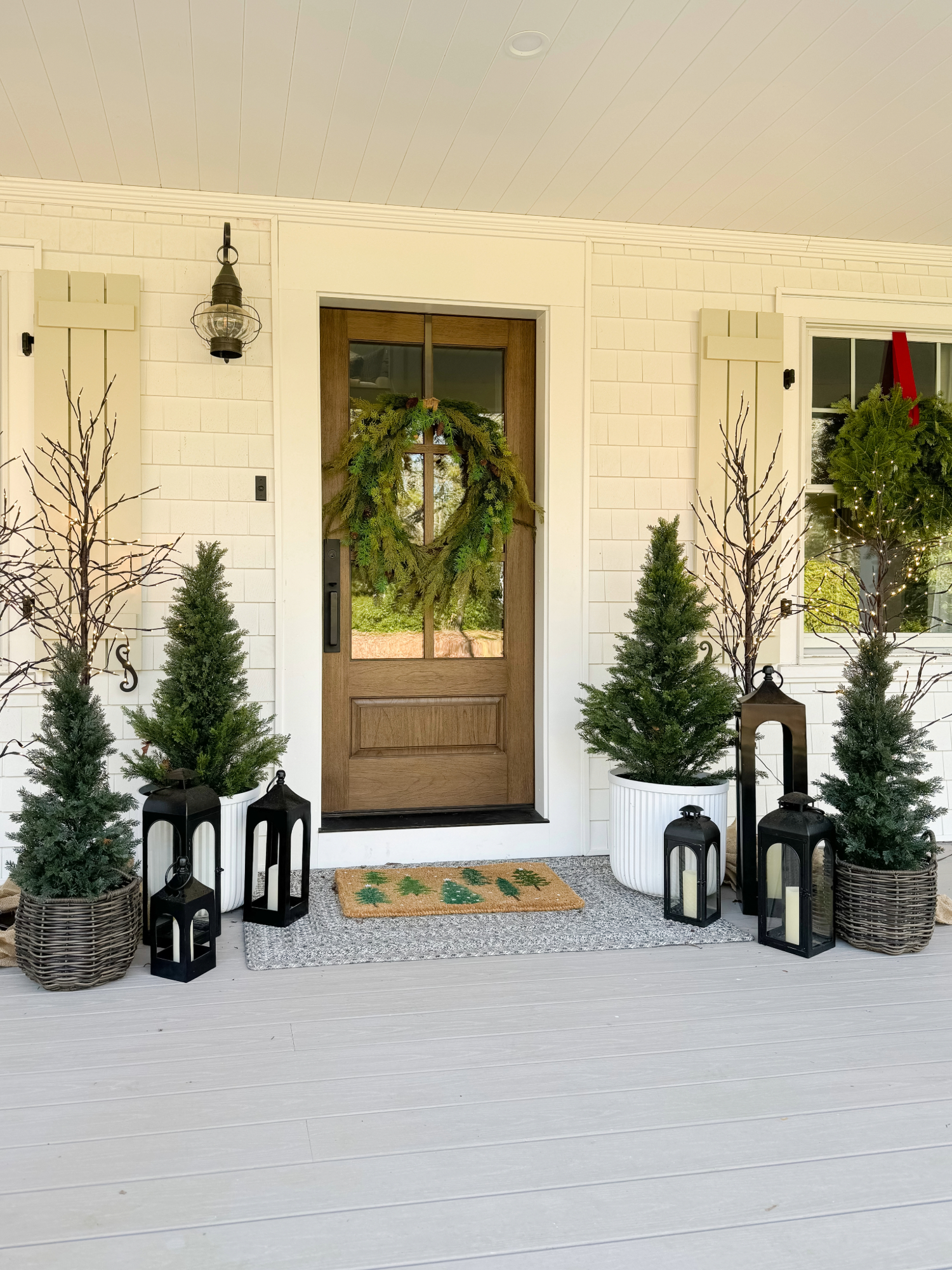 Coastal farmhouse front porch with natural wood door, fresh greenery wreath, potted evergreens, black lanterns, and woven baskets. Seasonal Amazon Prime Day home decor finds.