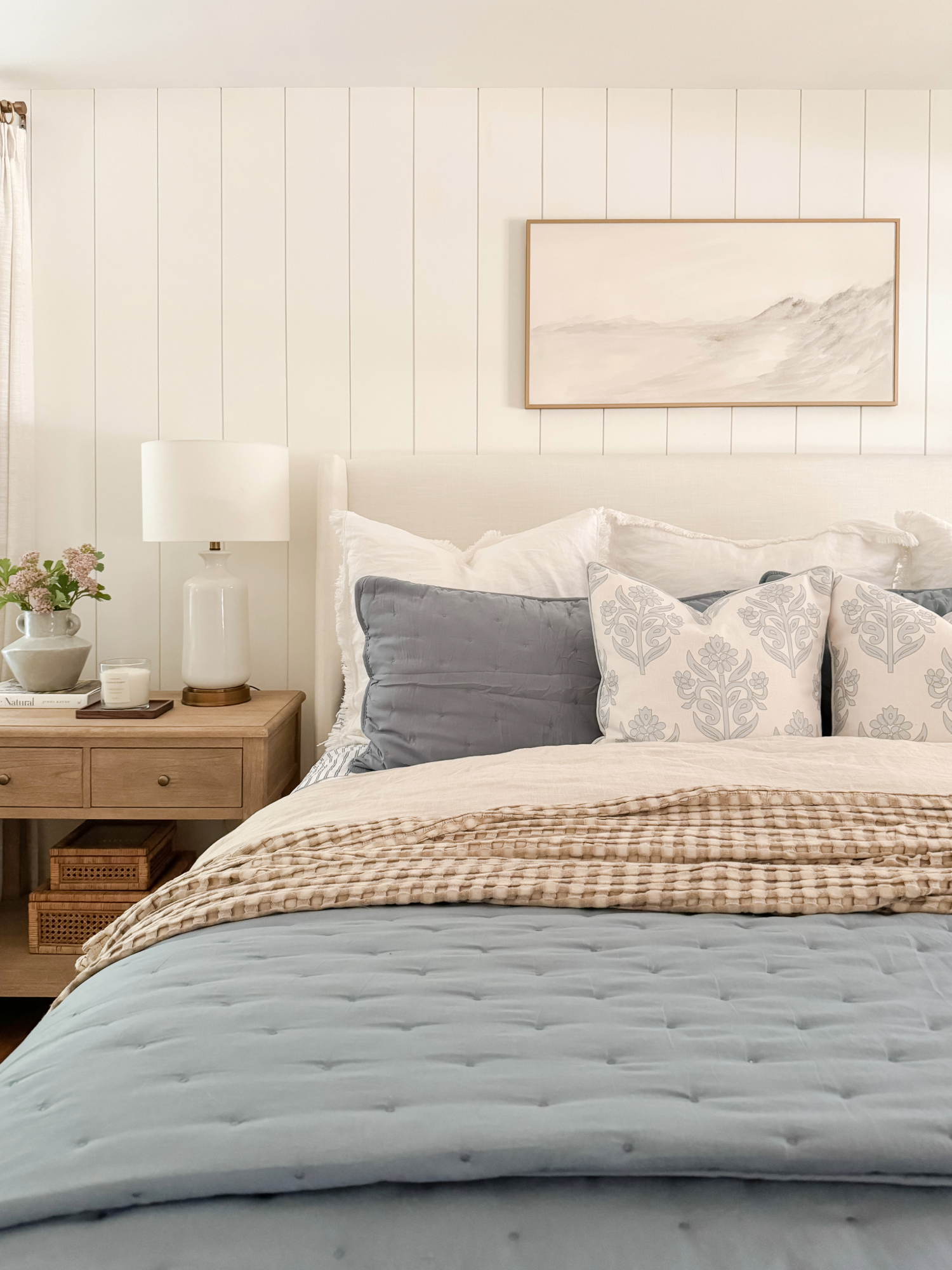 Coastal bedroom with layered blue and beige bedding, floral pillows, shiplap wall, and neutral nightstand decor. Coastal Amazon Prime Day home decor inspiration.