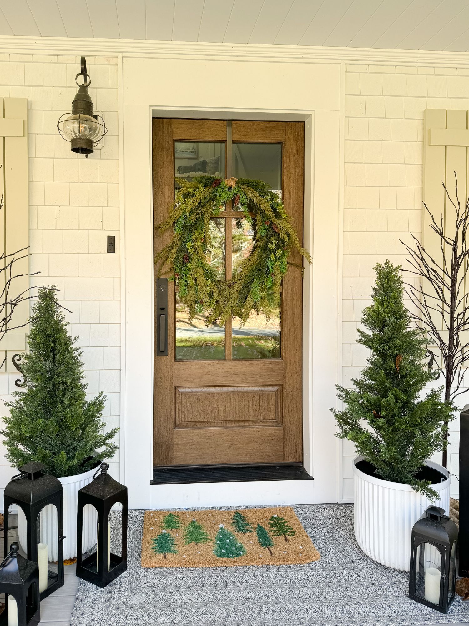 Front porch decorated for Christmas with a wood door, fresh evergreen wreath, lanterns, and potted trees for a classic holiday welcome.