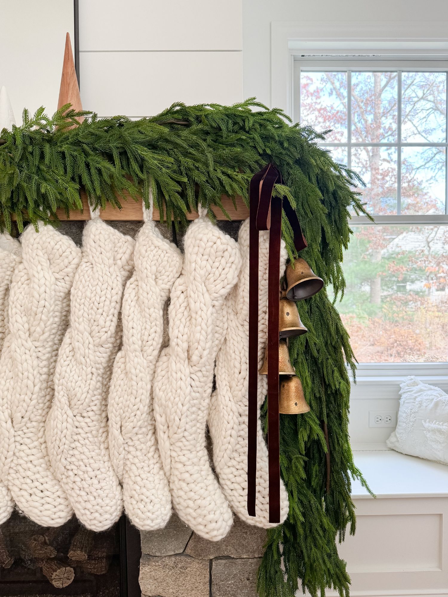Living room mantel decorated with fresh garland, knit stockings, and brass bells for a classic and cozy Christmas scene.