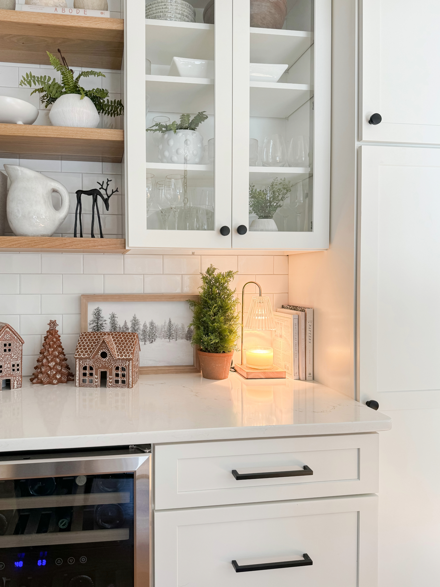 Neutral holiday kitchen styling with white cabinetry, glass cabinet doors, mini potted Christmas tree, gingerbread houses, and a glowing candle for a cozy seasonal touch.