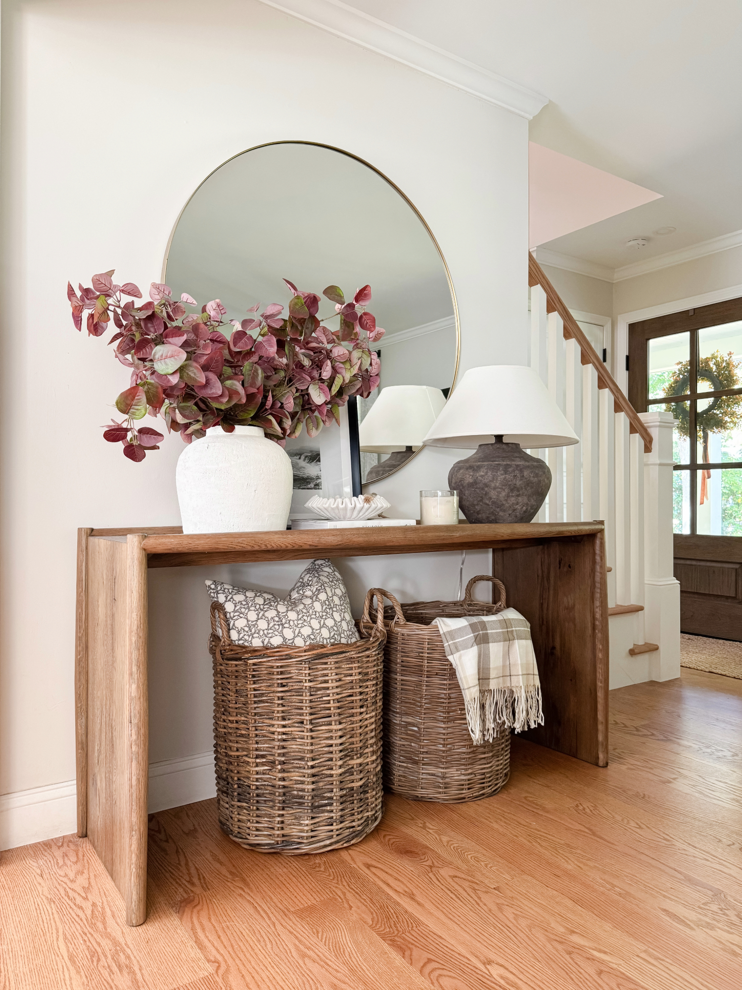 Warm neutral entryway with wooden console table, round gold mirror, wicker baskets, ceramic vases, and soft fall foliage for welcoming seasonal décor.