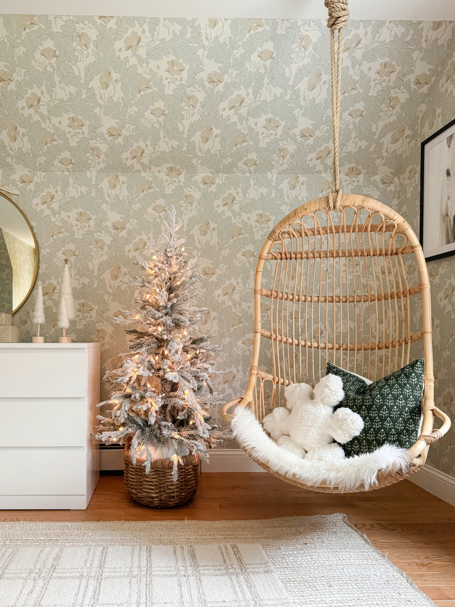 Cozy reading nook with rattan hanging chair, green throw pillows, faux fur cushion, and small flocked Christmas tree in a woven basket for warm winter décor inspiration.