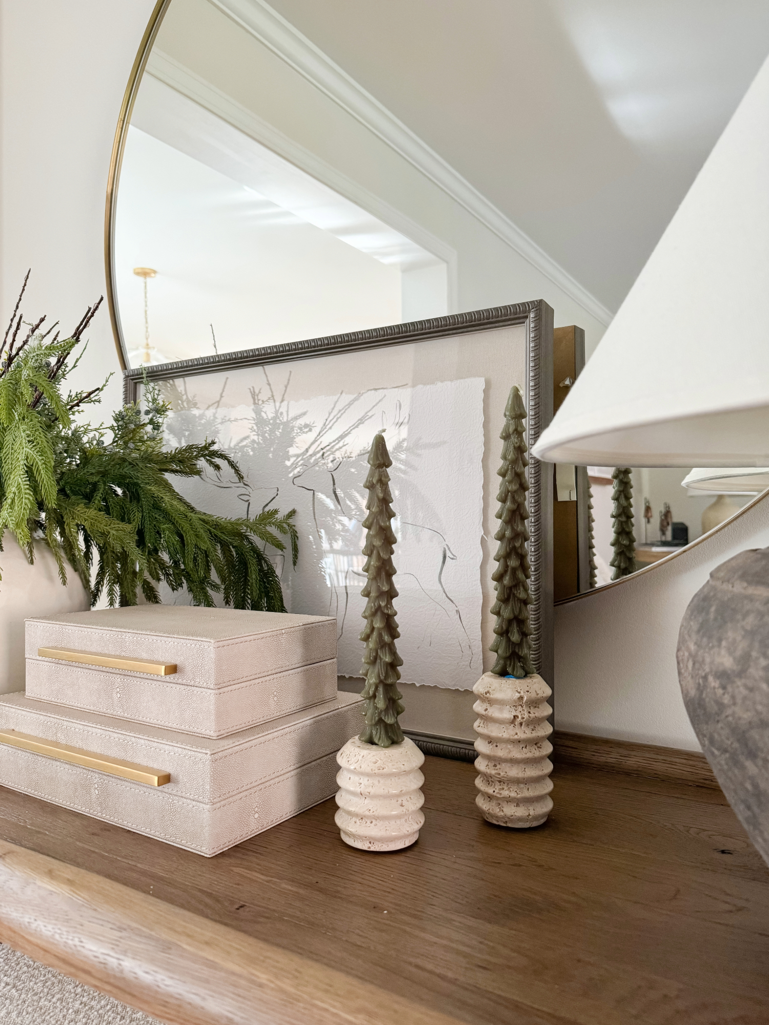 Minimalist console table styled with modern artwork, evergreen branches, textured boxes with brass handles, and ceramic Christmas tree décor for an elegant holiday entryway.