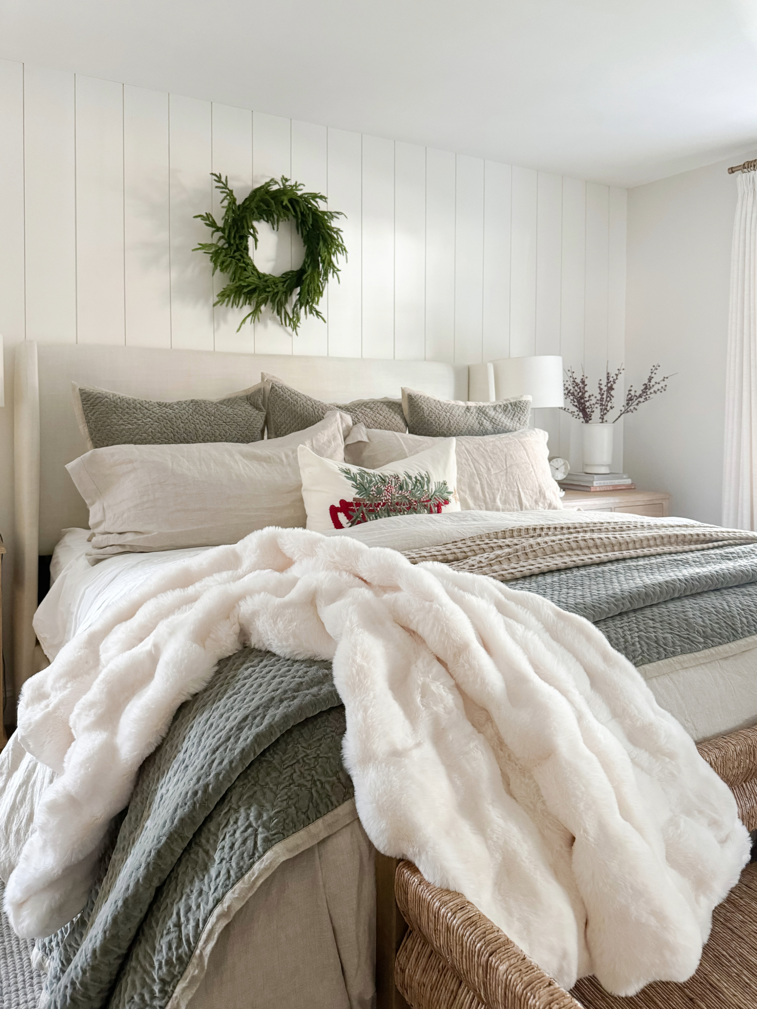 Cozy winter bedroom with layered bedding, soft faux fur throw, neutral linens, and a greenery wreath for simple and timeless holiday bedroom décor.