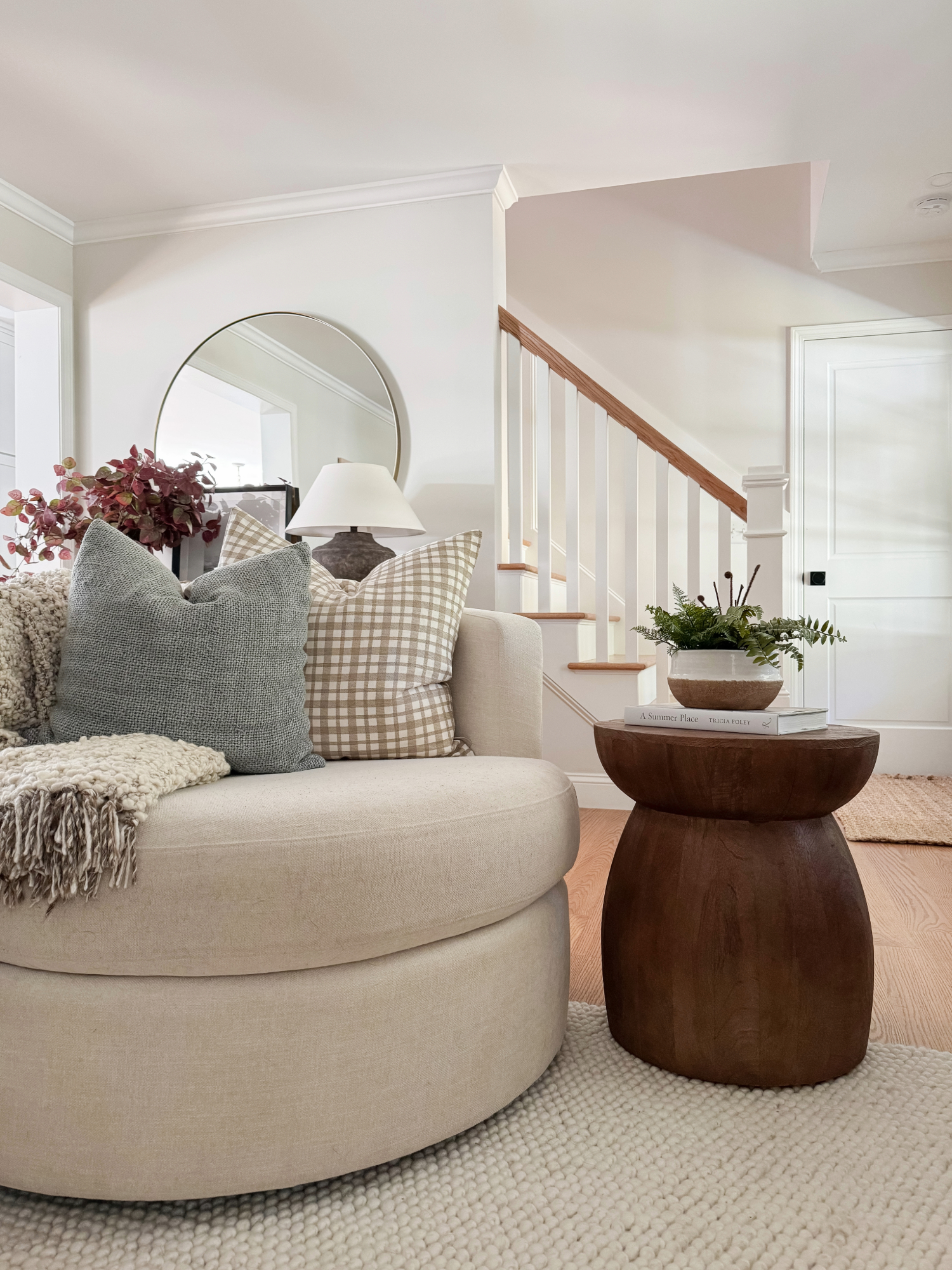 Neutral living room corner featuring a round swivel chair, textured throw pillows, knit blanket, and wood accent table with fall greenery and stacked books for cozy seasonal style.