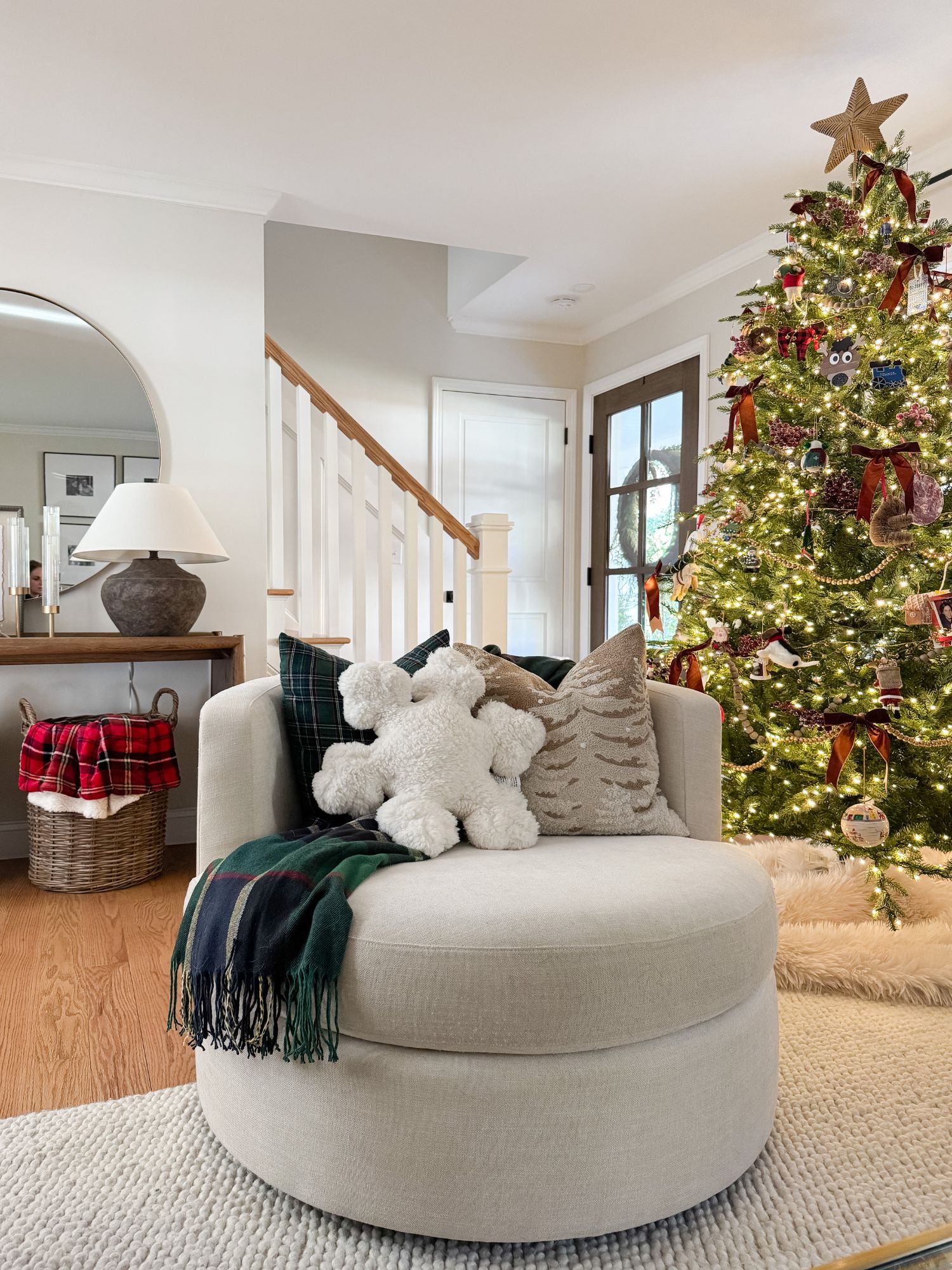Cozy living room corner with a neutral swivel chair, plaid throw blanket, snowflake pillow, and a decorated Christmas tree glowing beside a console table and round mirror.