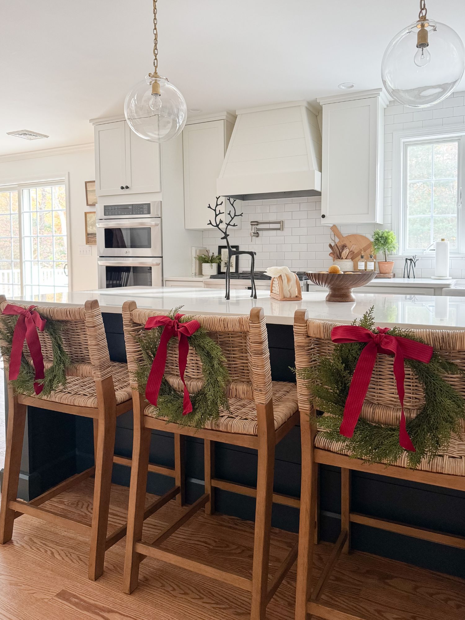 Bright kitchen with three woven counter stools decorated with small wreaths and red bows, a large island, clear globe pendant lights, and white cabinetry.