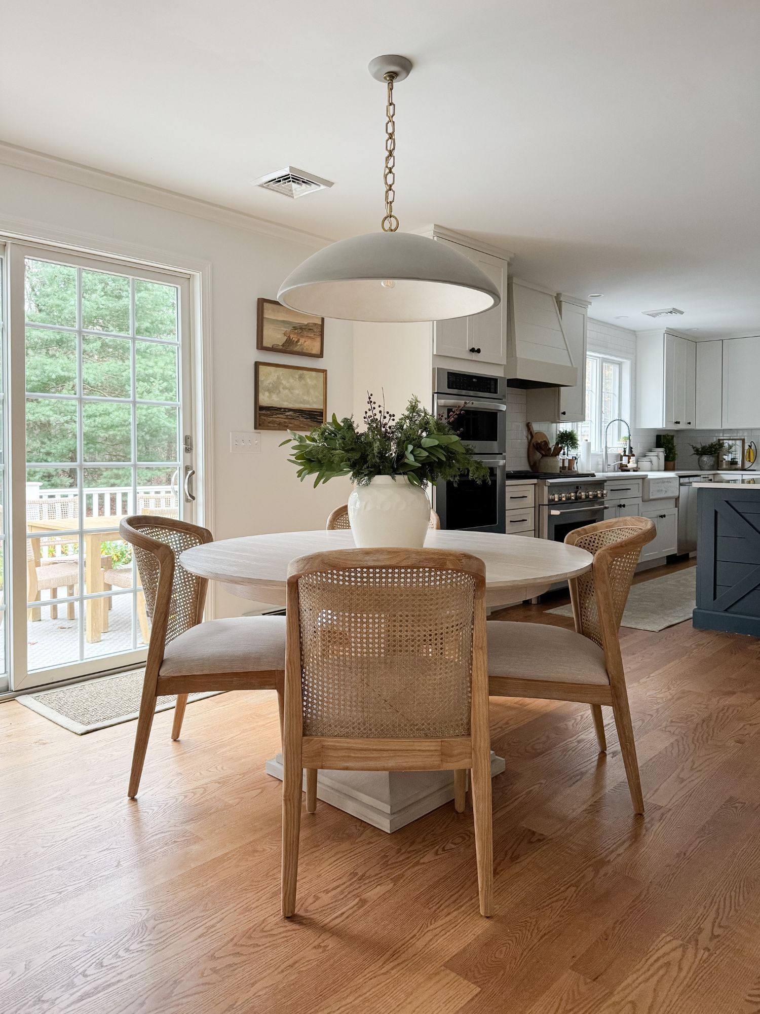 Dining space with a round wooden table, four cane dining chairs, a large dome pendant light, and a vase of greenery as the centerpiece.