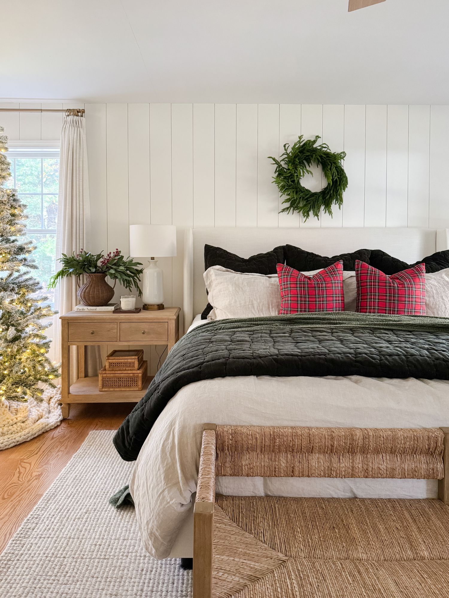Holiday bedroom with an upholstered bed, plaid pillows, a green quilt, woven bench, Christmas tree, nightstand with a lamp, and a green wreath above the bed.