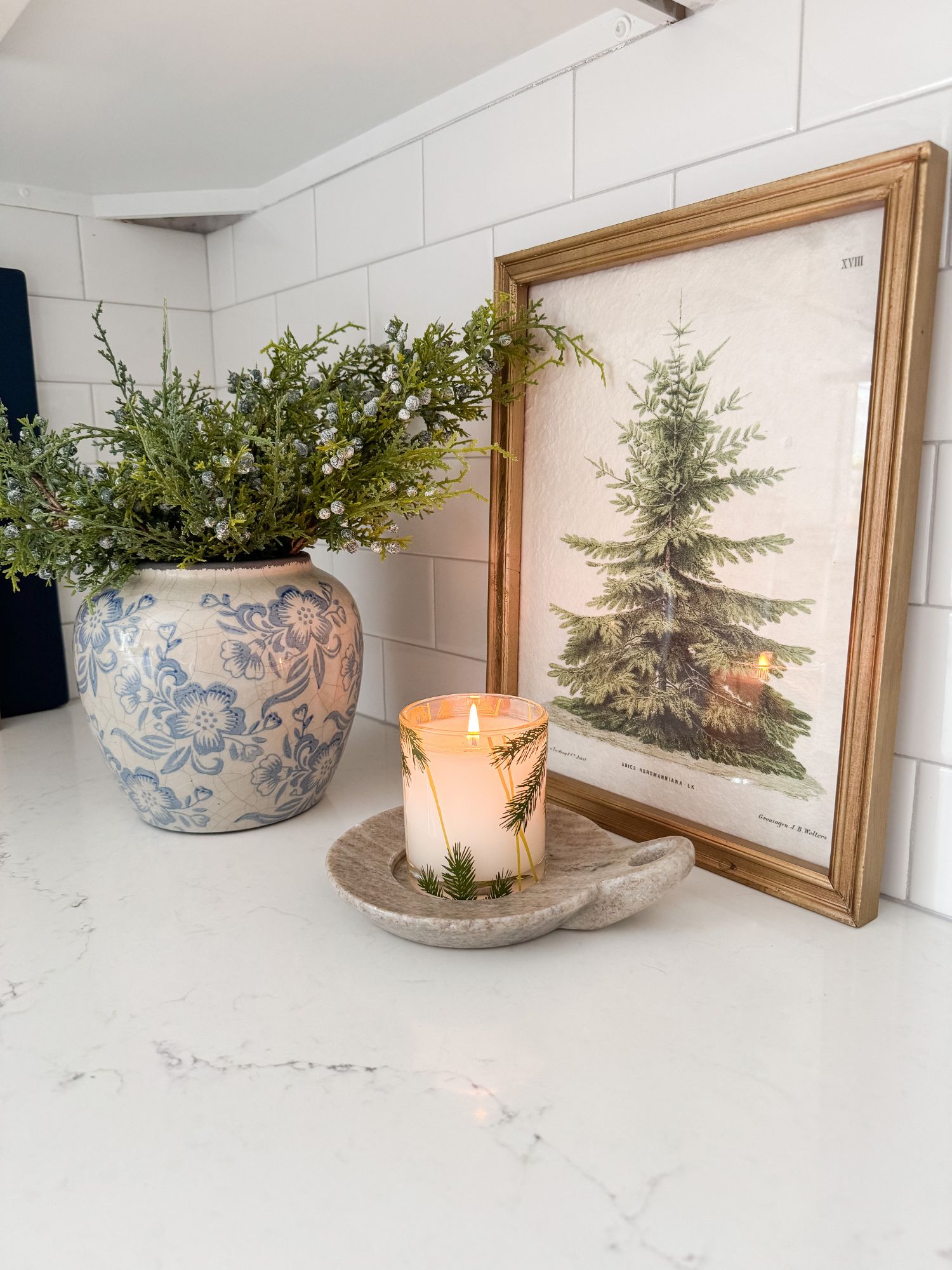 Kitchen counter vignette featuring a blue floral vase with winter greenery, a framed evergreen print, and a holiday candle on a stone dish against white tile backsplash.