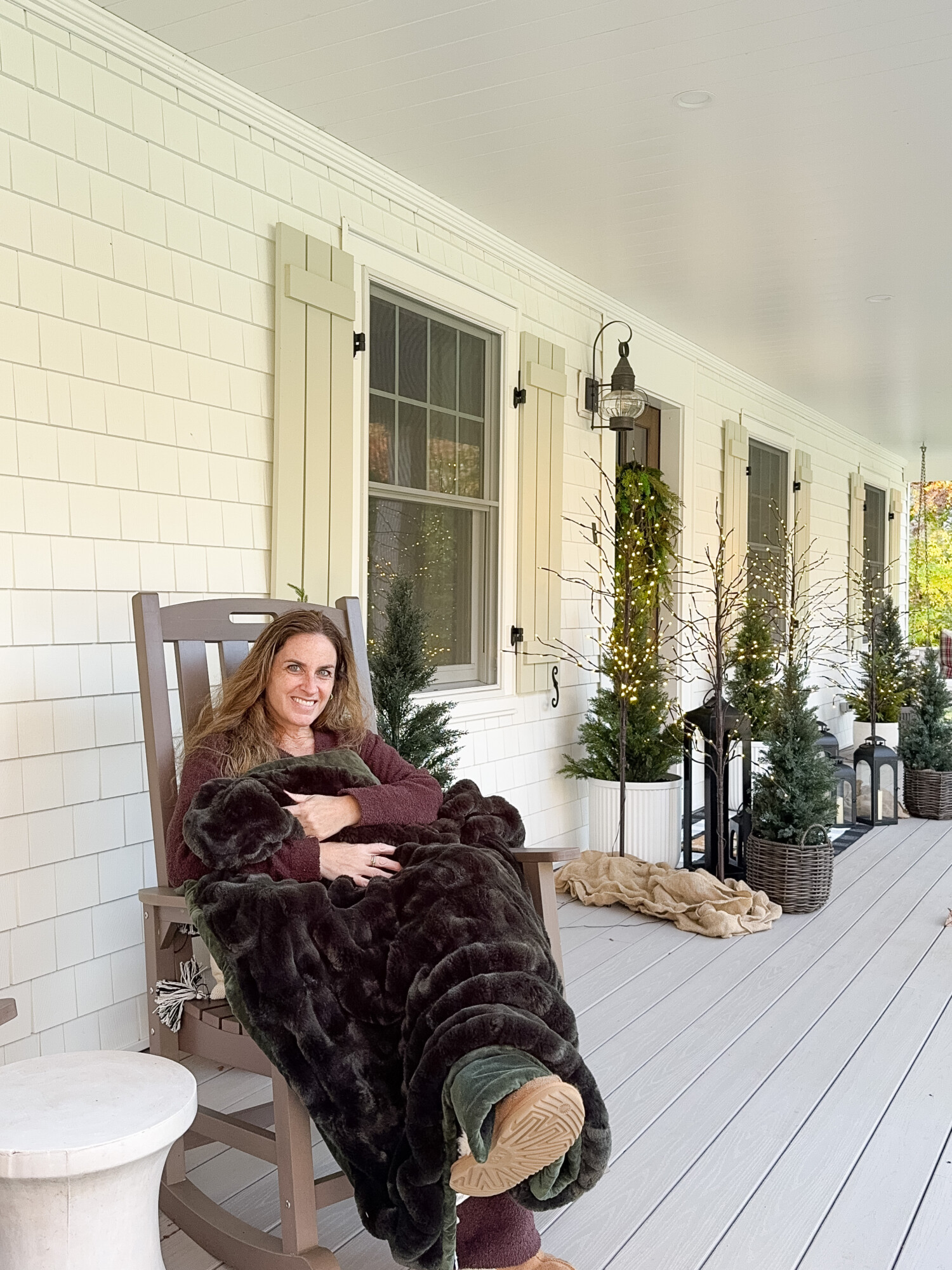 Sherri Calnan sitting on a front porch in a rocking chair, wrapped in a dark green faux fur blanket and wearing cozy loungewear and slippers. The porch is decorated with potted evergreens and twinkling lights, creating a warm and inviting holiday atmosphere.