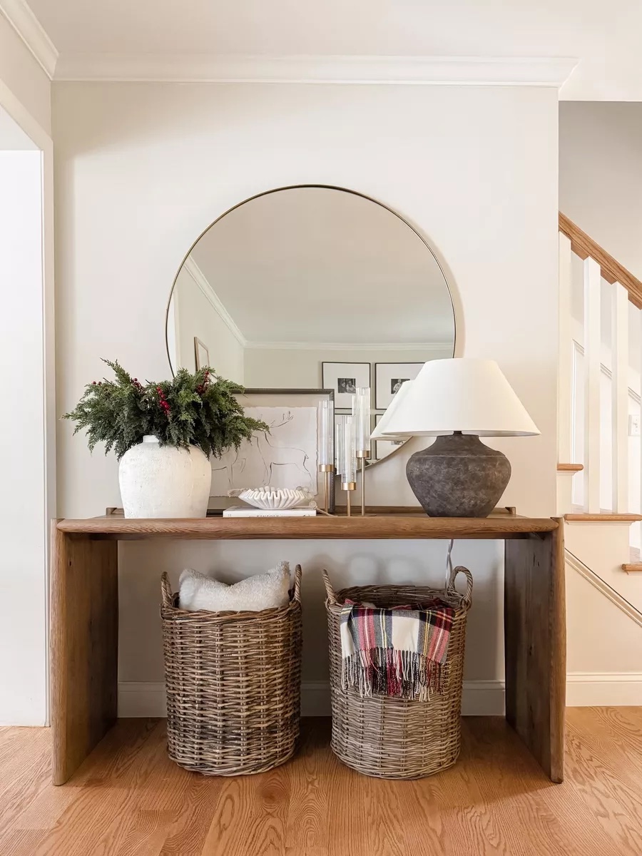 Entryway console table styled with a round gold mirror, table lamp, winter greenery in a white vase, candles, and woven baskets filled with pillows and blankets.