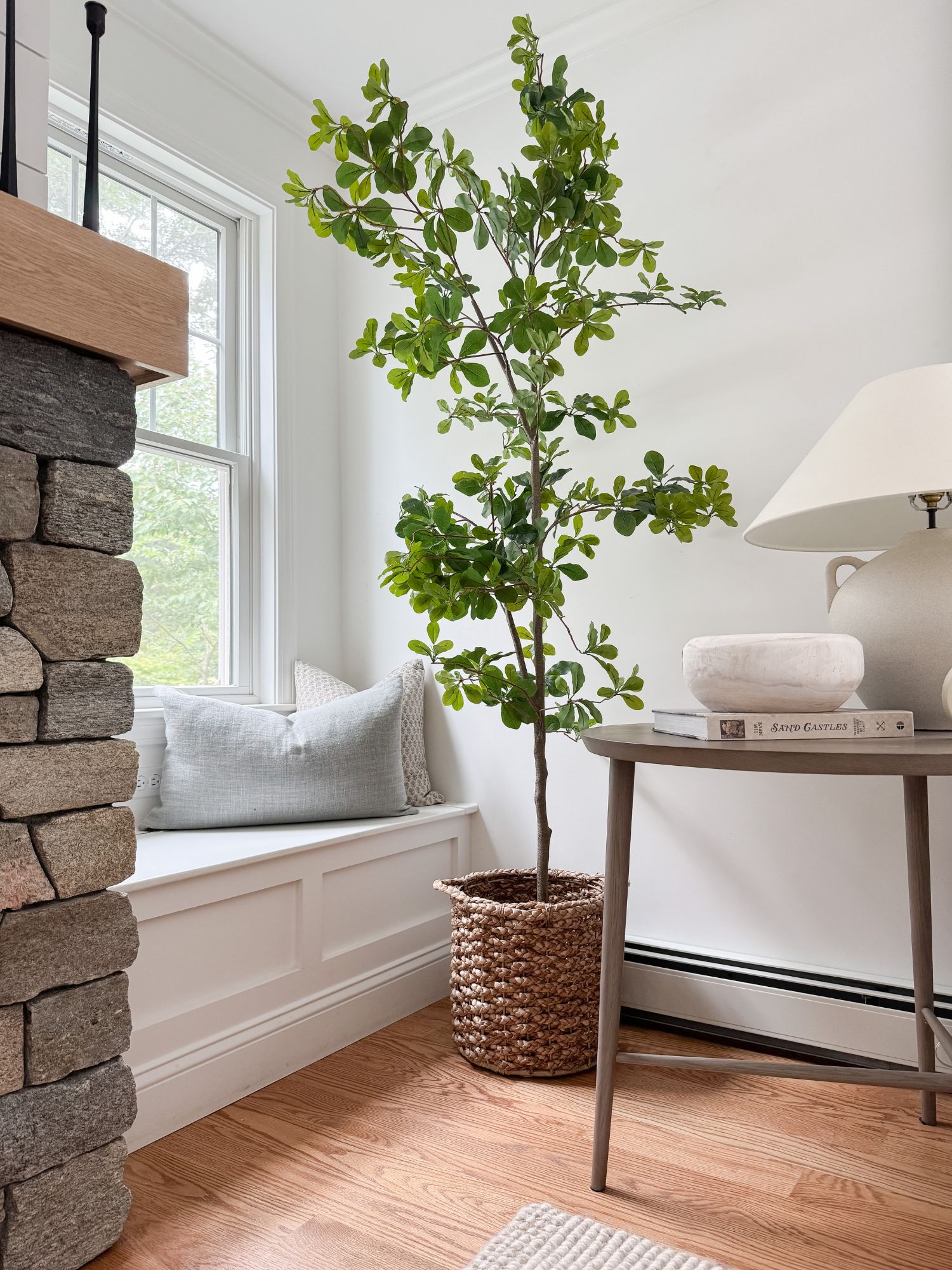 Coastal corner with a tall faux tree placed in a woven basket beside a window seat and neutral decor