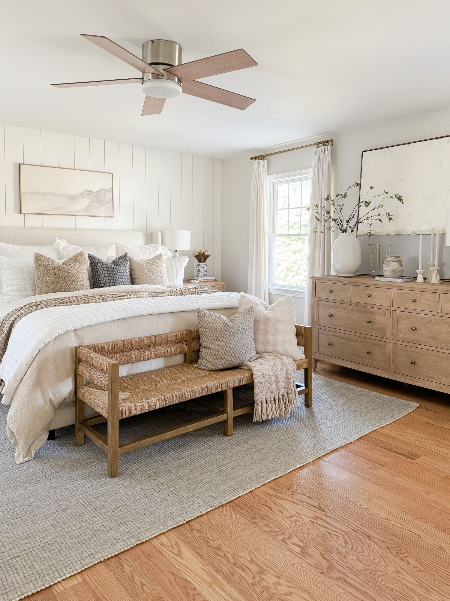 Coastal bedroom with an upholstered bed, layered neutral bedding, woven bench, wood dresser, and light wood floors