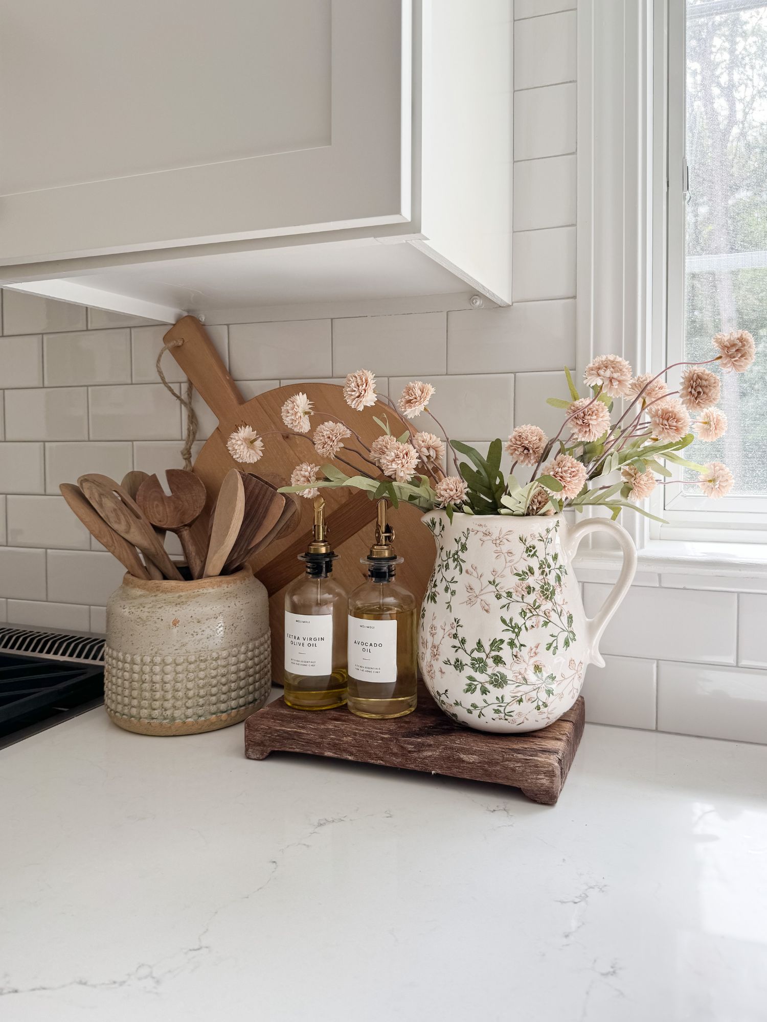Coastal kitchen countertop styled with oil and vinegar dispensers, wooden cutting boards, ceramic utensil holder, and soft neutral florals