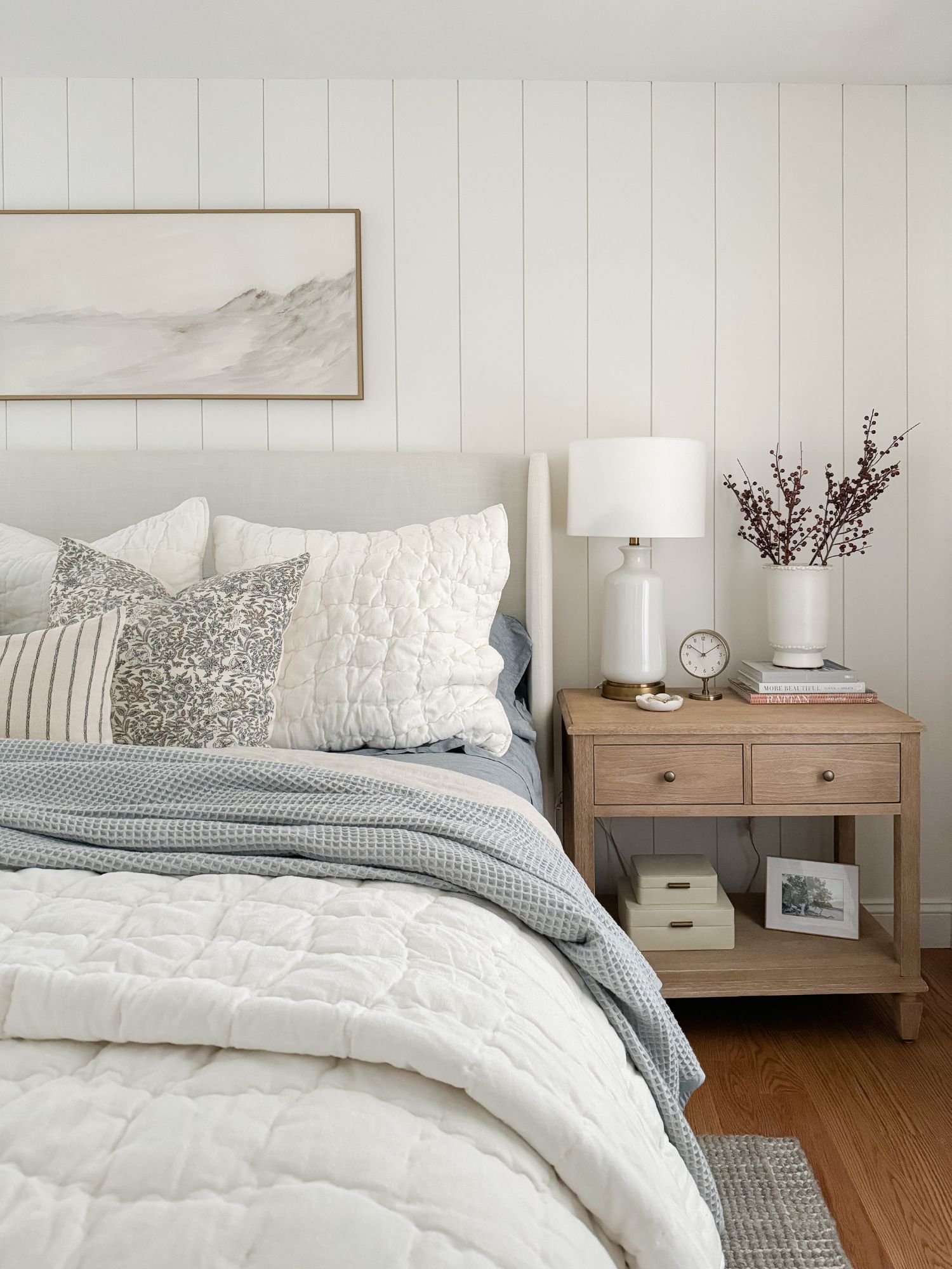 Winter bedroom nightstand styled with white ceramic lamp, clock, neutral books, and simple berry branches.