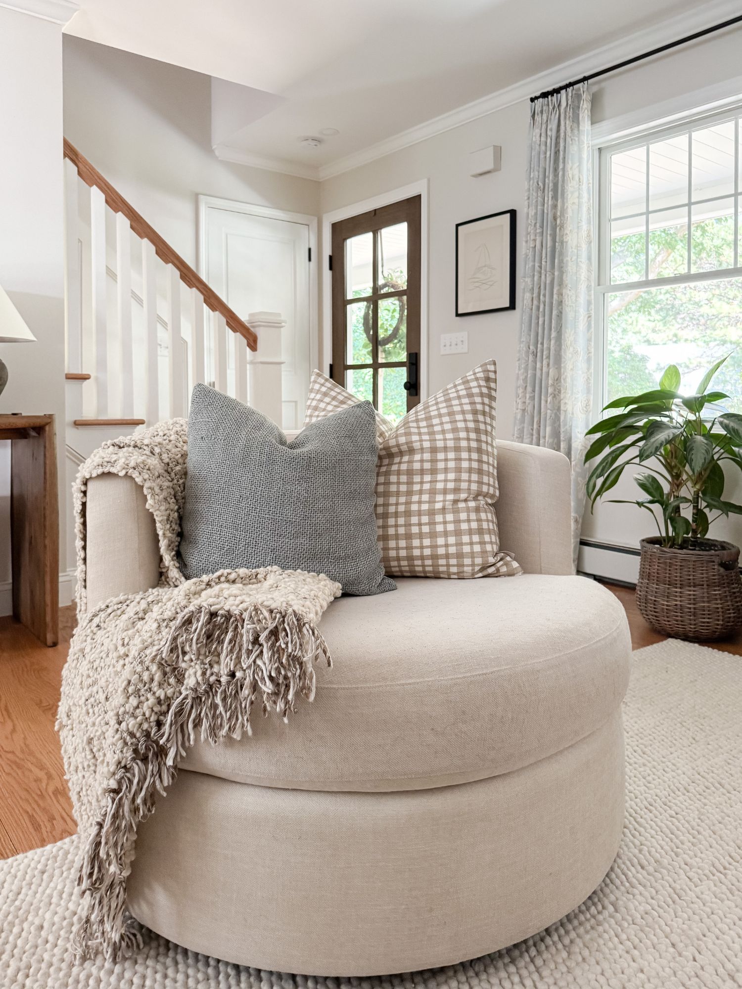 Neutral living room corner with a round upholstered swivel chair, textured throw, plaid pillows, and natural light