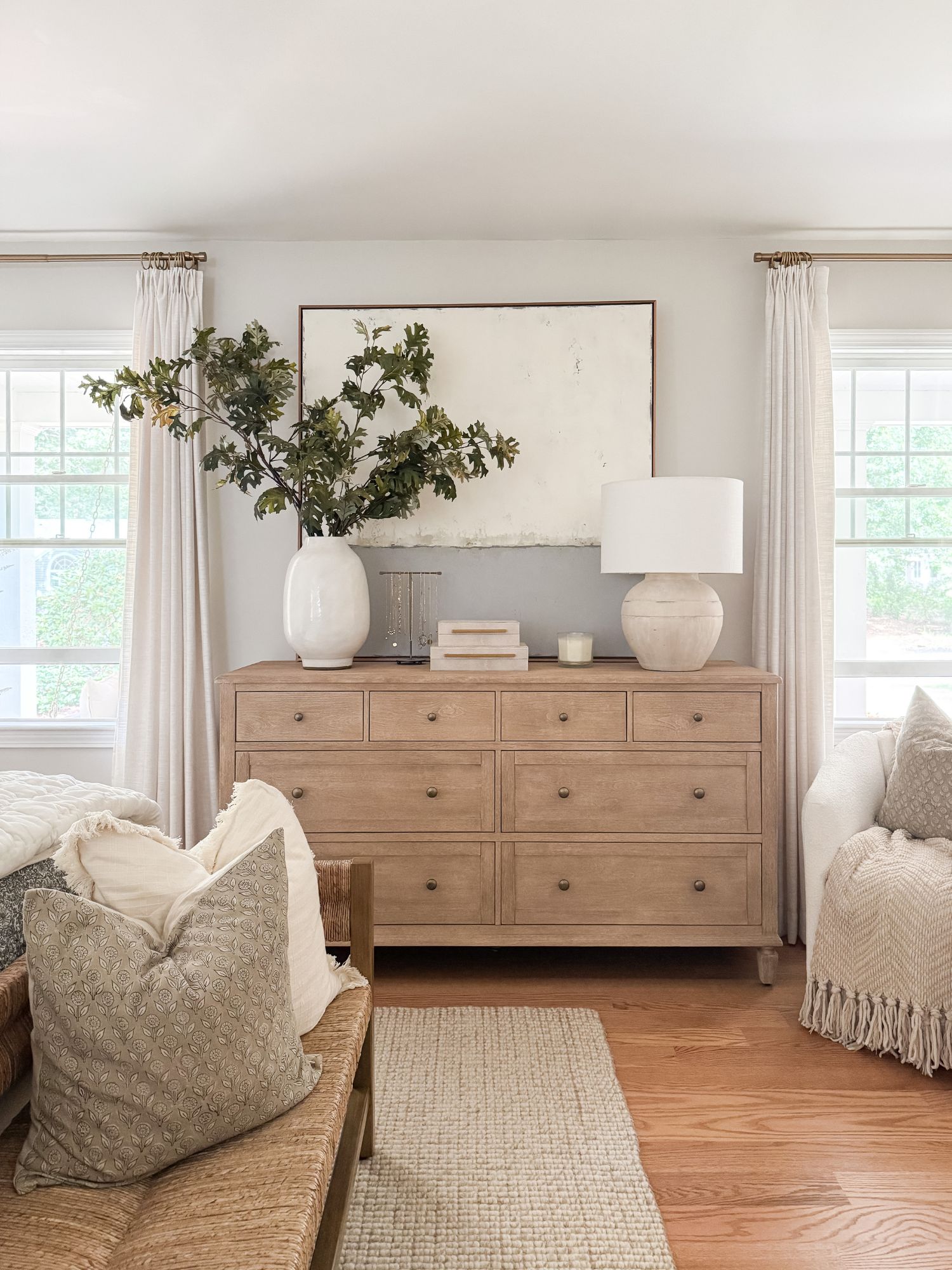 Light wood dresser styled with ceramic lamp, large neutral artwork, books, and greenery for a coastal bedroom look