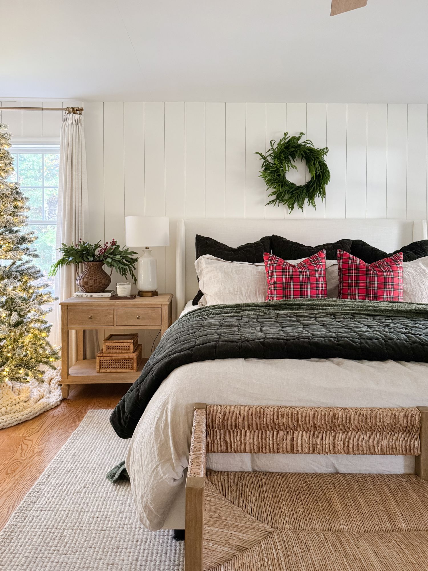 Christmas winter bedroom with layered white bedding, deep green throw, woven bench, and soft holiday lighting from the tree.