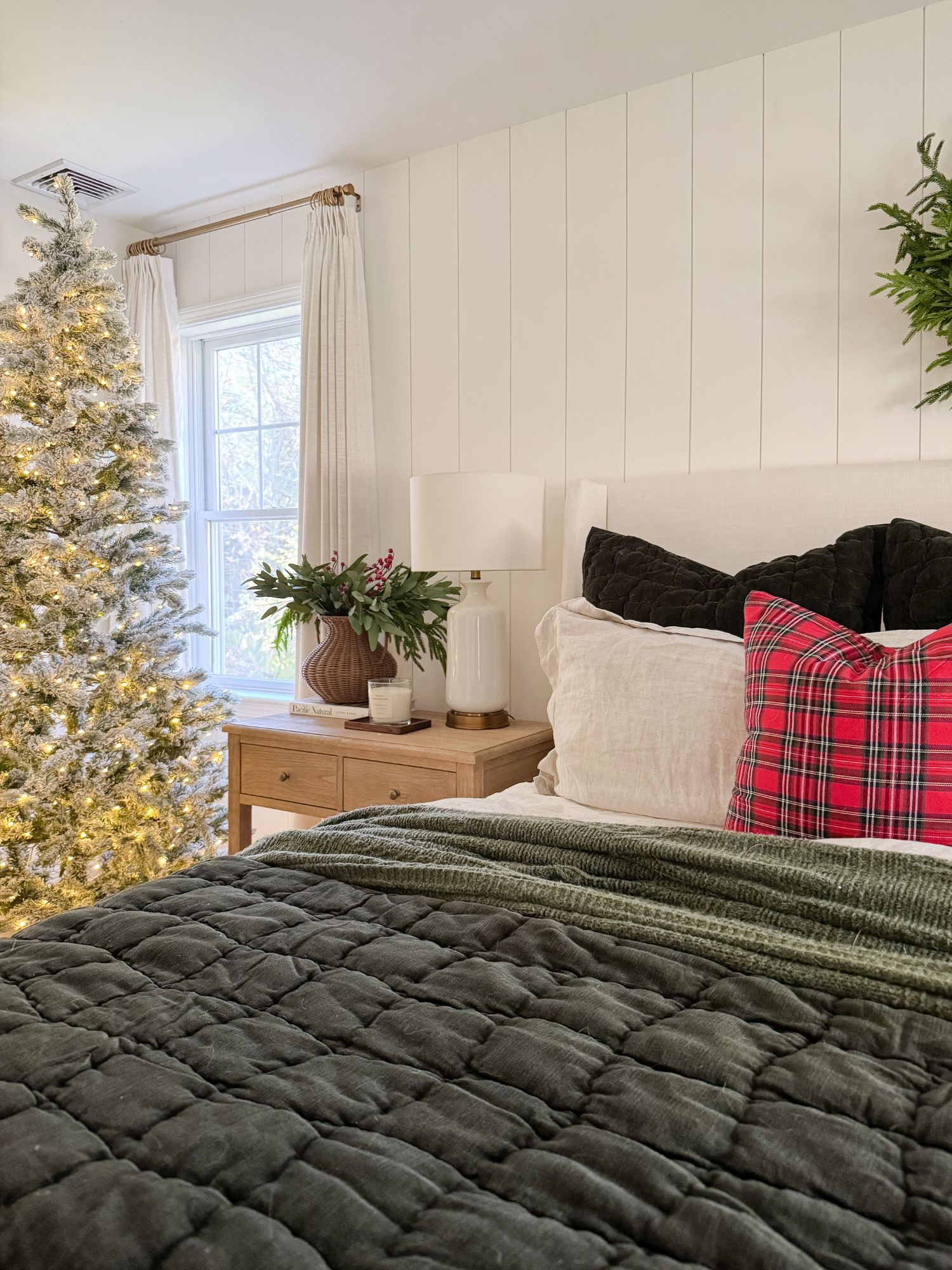 Winter bedroom styled for Christmas with green quilt, red plaid pillows, natural wreath, and flocked Christmas tree beside the bed.