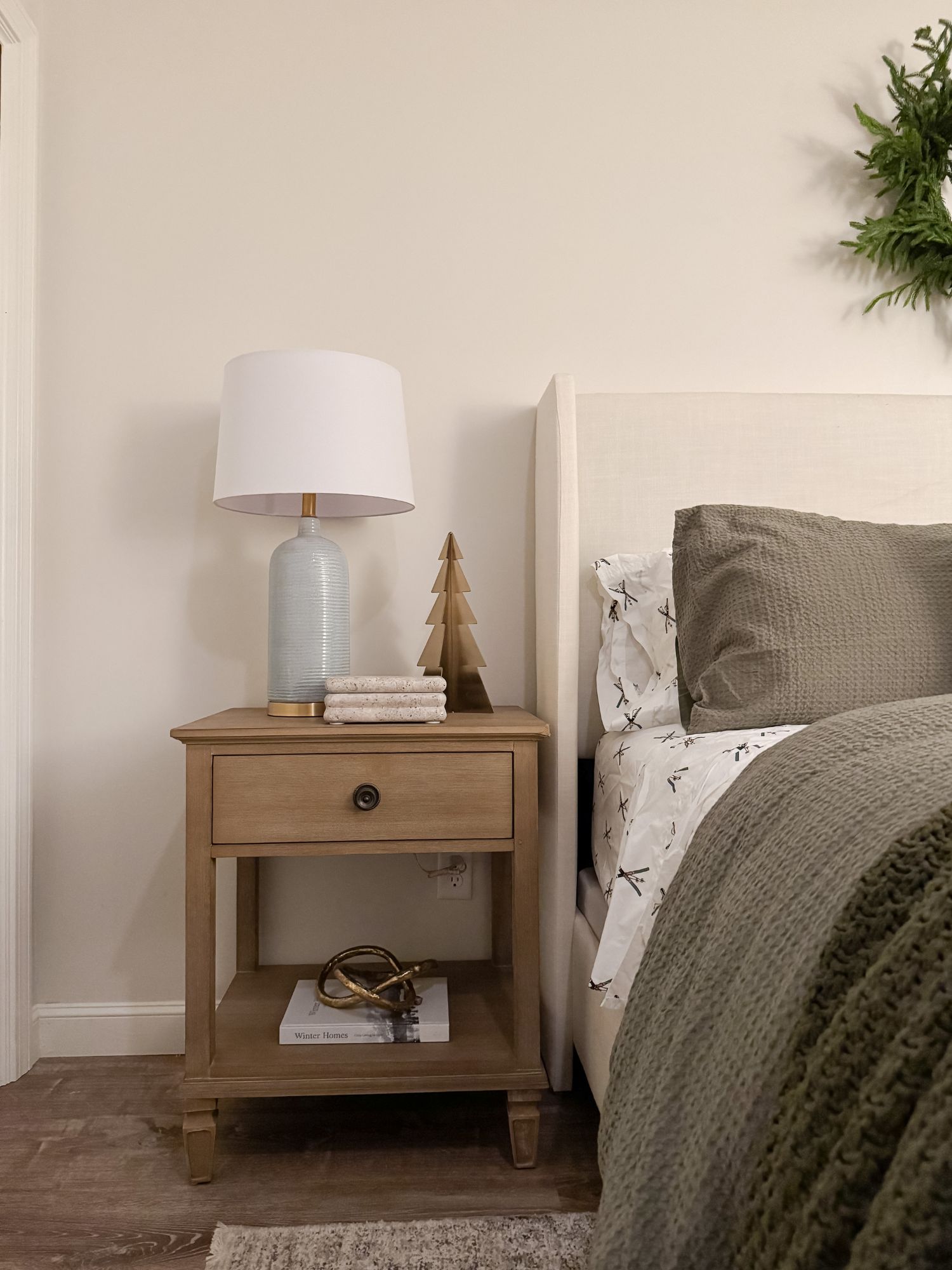 Holiday winter bedroom nightstand with blue glass lamp, brass tree decor, stacked books, and neutral bedding.