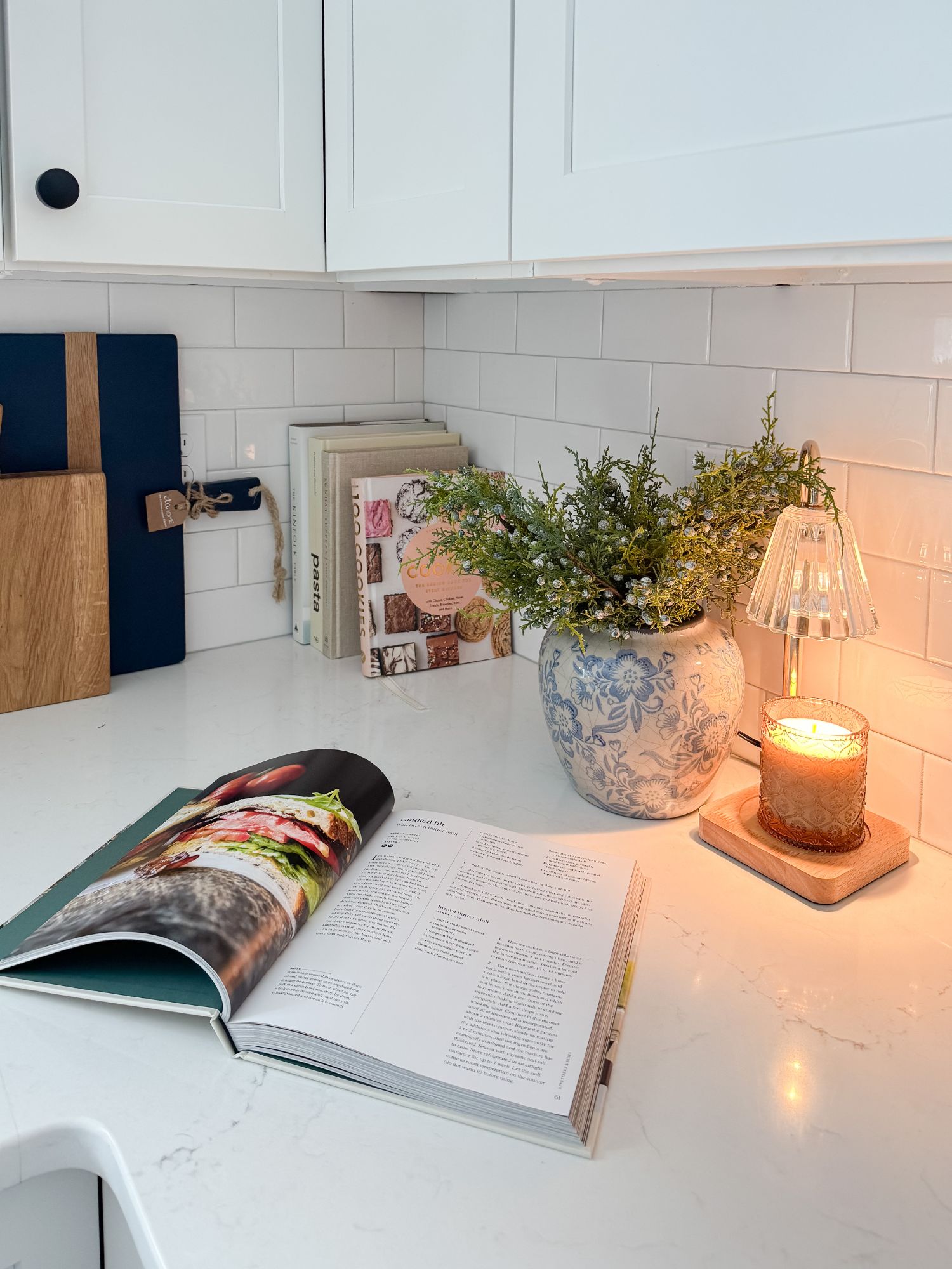 Cape Cod kitchen counter styled with a ceramic vase, winter greenery stems, candle warmer with cozy scent, and neutral cookbooks for winter decor.