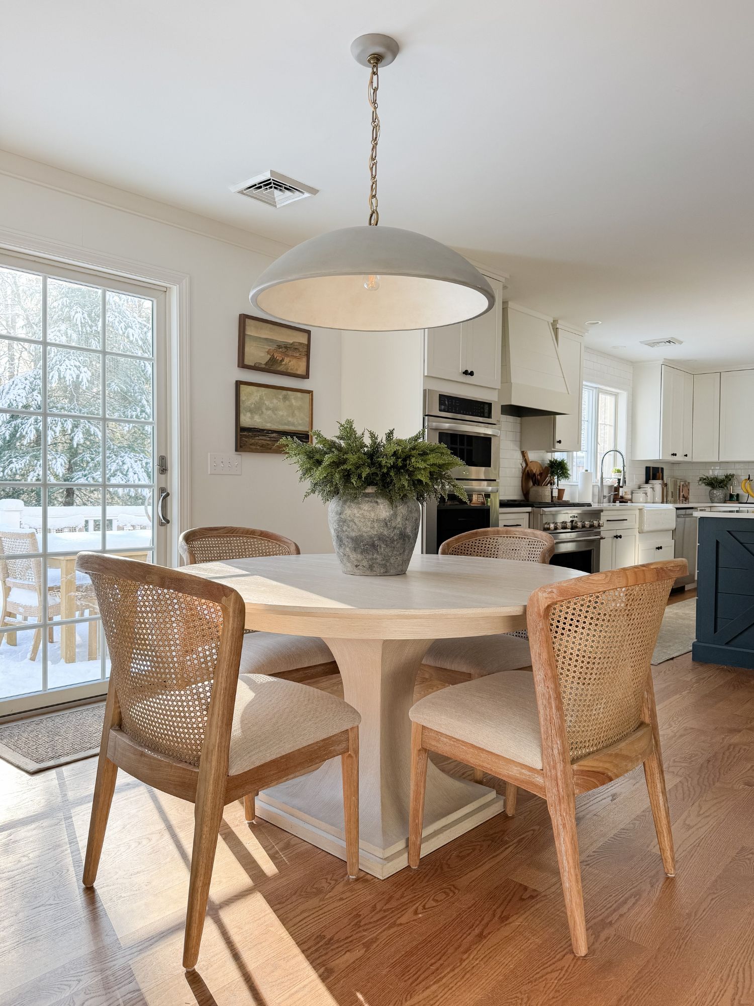 Cape Cod inspired kitchen dining area with rattan chairs, round wood table, winter greenery centerpiece, and soft neutral finishes.