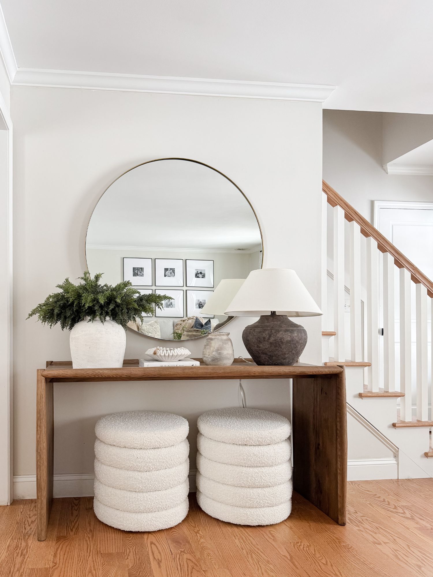 Cape Cod inspired console table styled with a round mirror, ceramic table lamp, winter greenery stems, and woven stools in a neutral entryway.