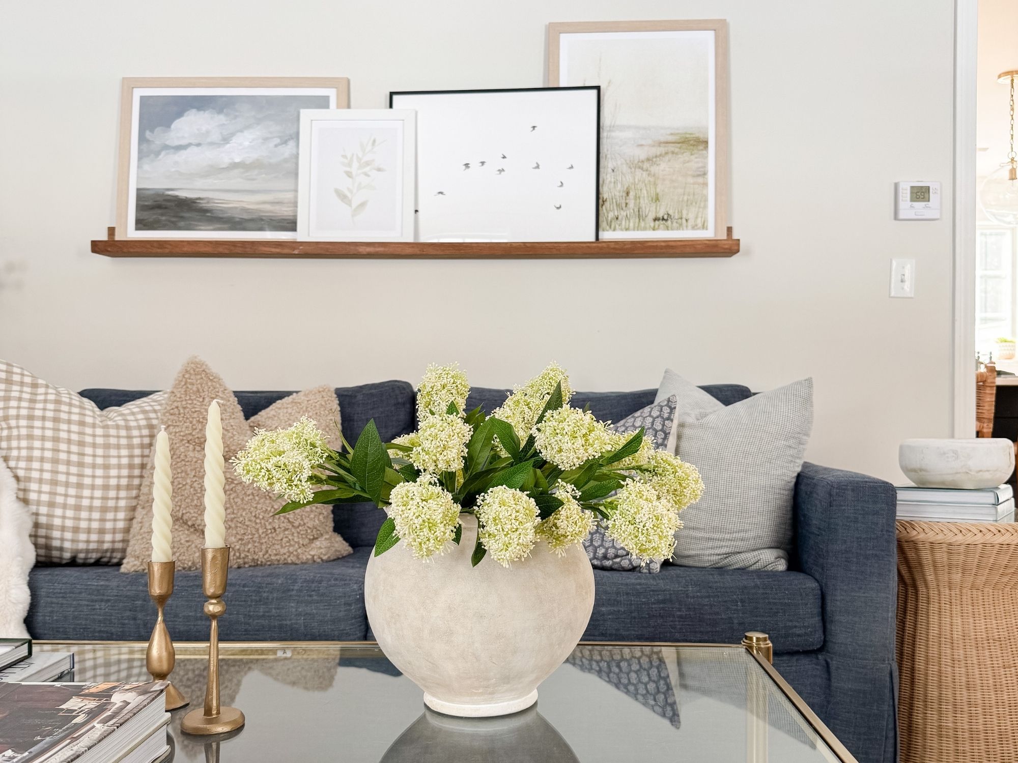Light and airy coastal living room with navy sofa, neutral pillows, ceramic vase with white spring flowers, brass candlesticks, and framed coastal artwork.