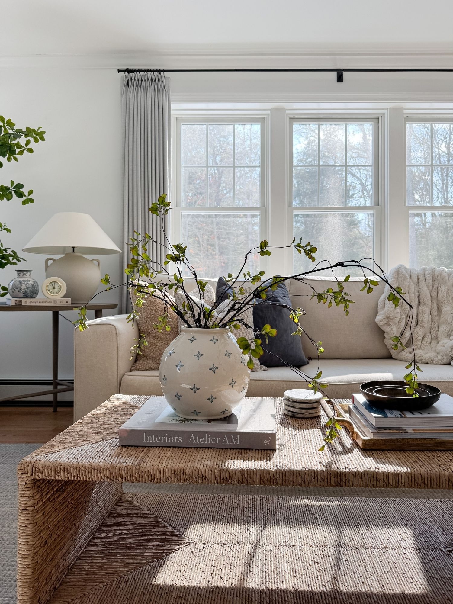 Coastal inspired family room with woven coffee table, ceramic vase with greenery branches, neutral sofa, linen curtains, and natural fiber rug.