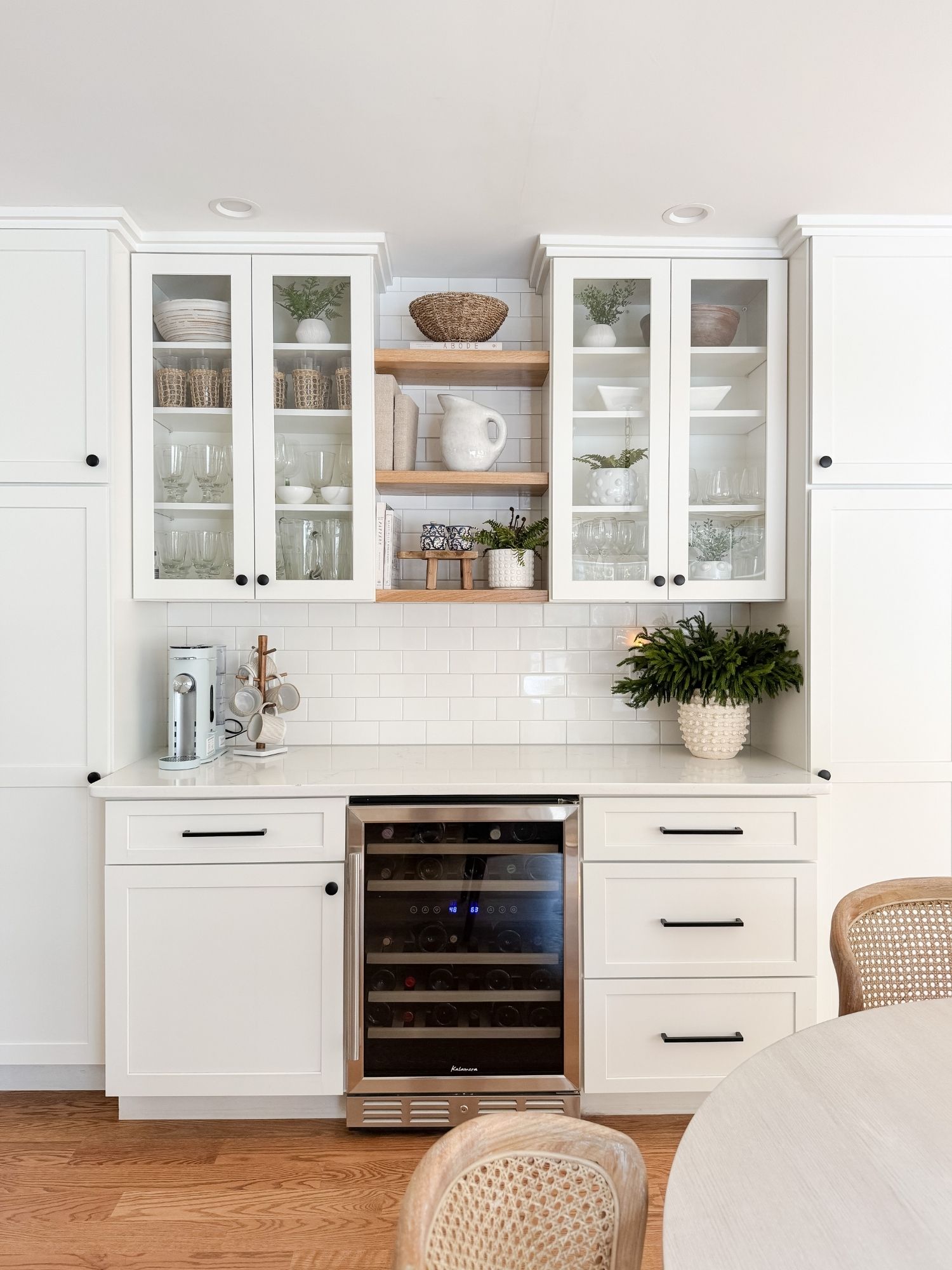Cape Cod inspired coffee bar styling with white ceramic pitcher, greenery, woven basket, and natural wood accents in a neutral coastal kitchen.