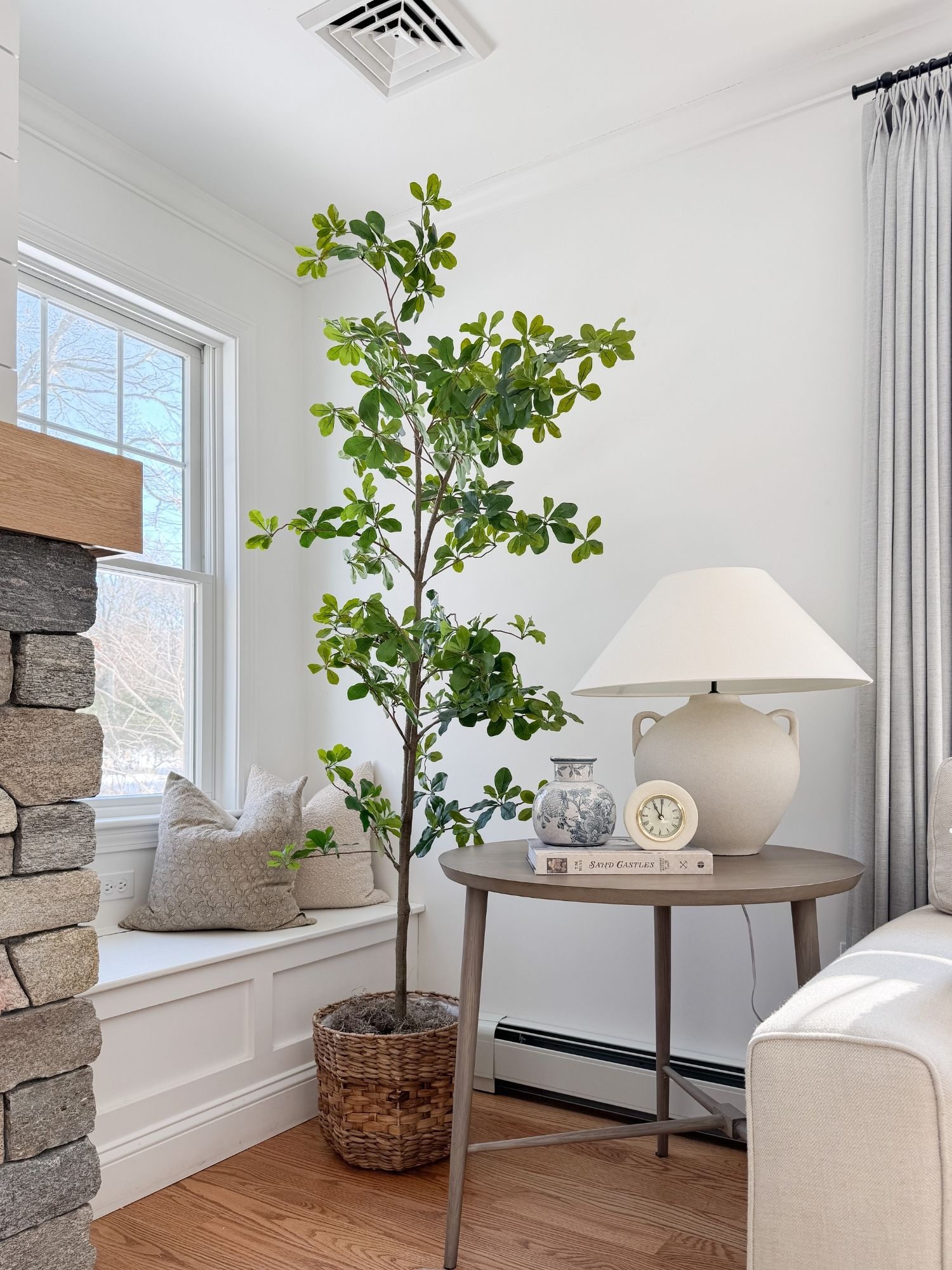 Neutral coastal living room corner with tall indoor tree in woven basket, ceramic lamp, soft pillows, and light filled windows.