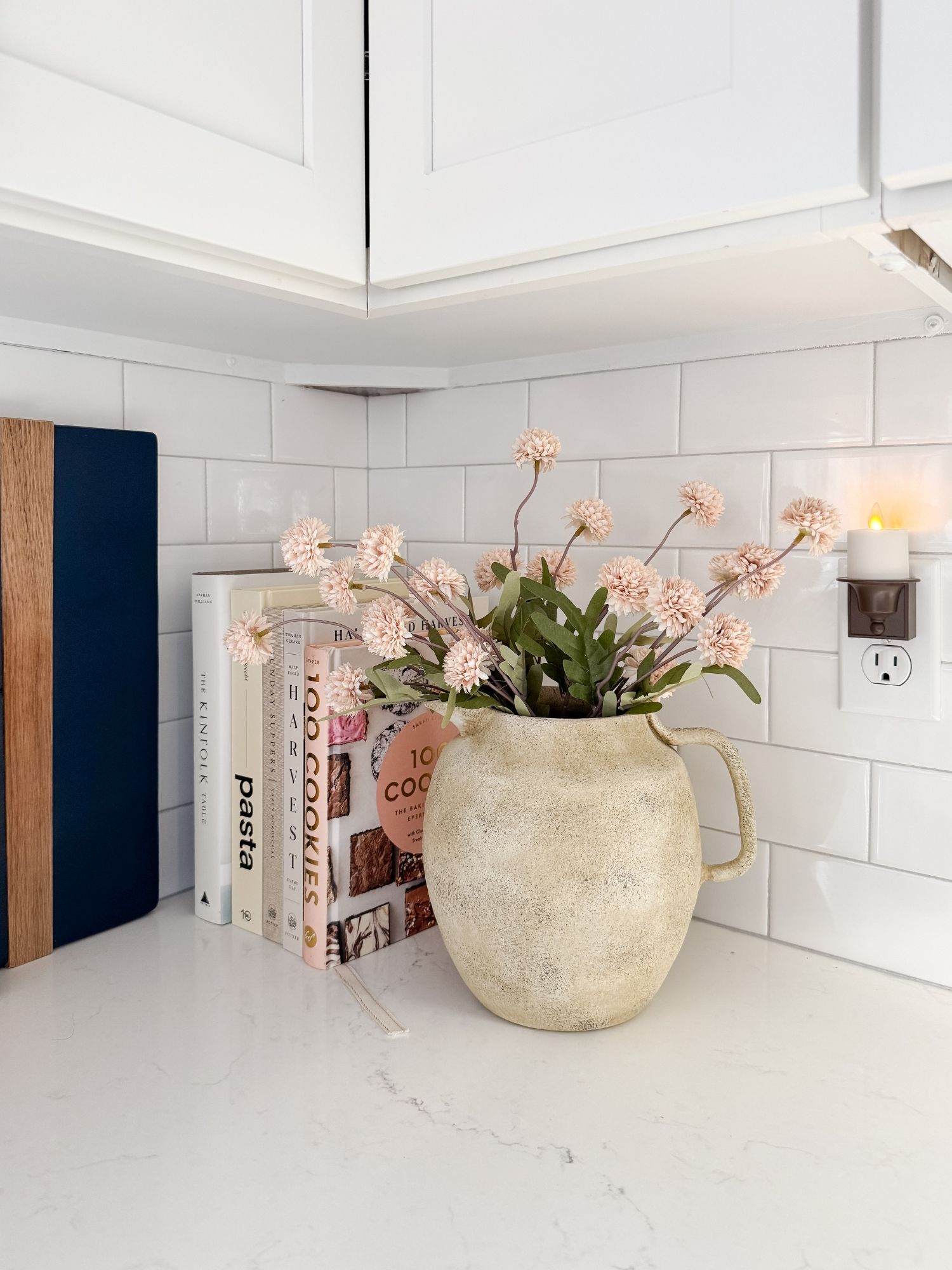 Coastal kitchen styling with textured ceramic pitcher, blush spring florals, stacked cookbooks, and white subway tile backsplash.