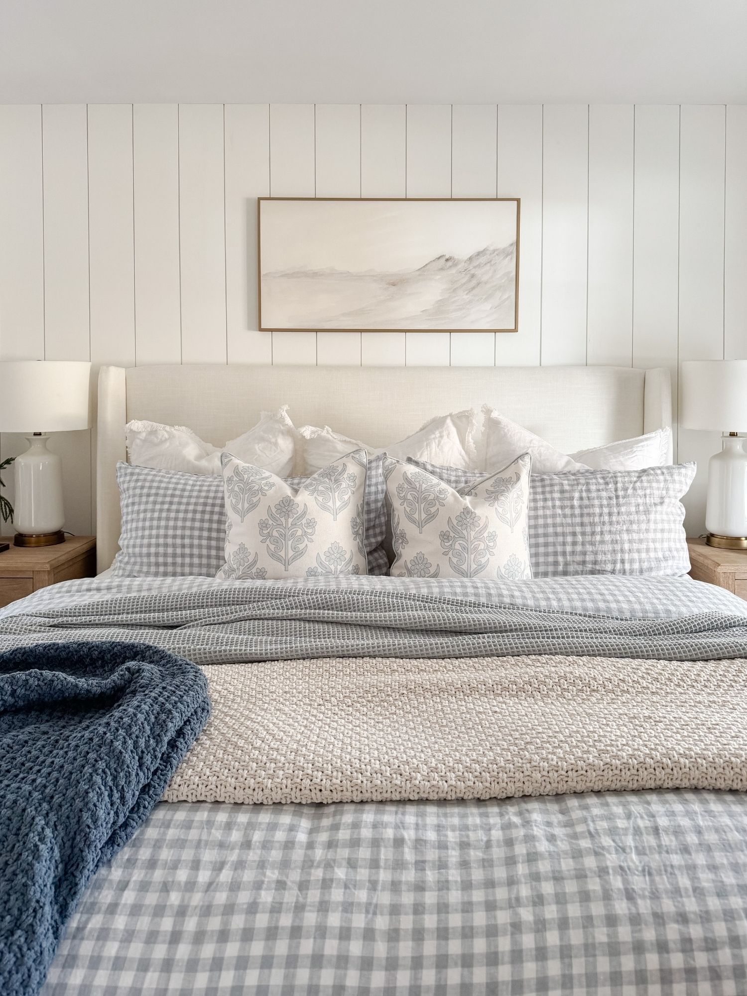 Serene coastal bedroom with light gingham bedding, layered neutral pillows, soft blue throw blanket, and neutral landscape artwork.