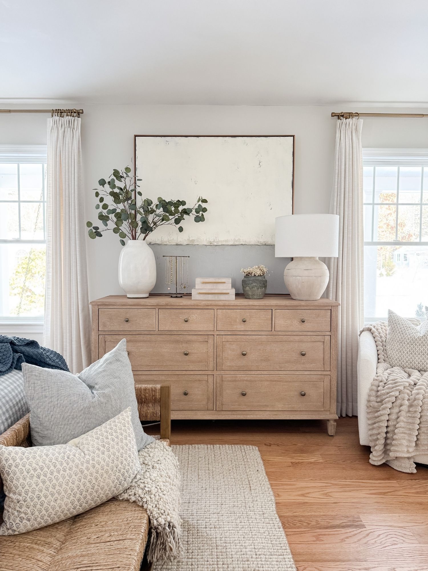 Cape Cod inspired bedroom with light wood dresser, ceramic lamp, neutral artwork, woven accents, and greenery in white vase.