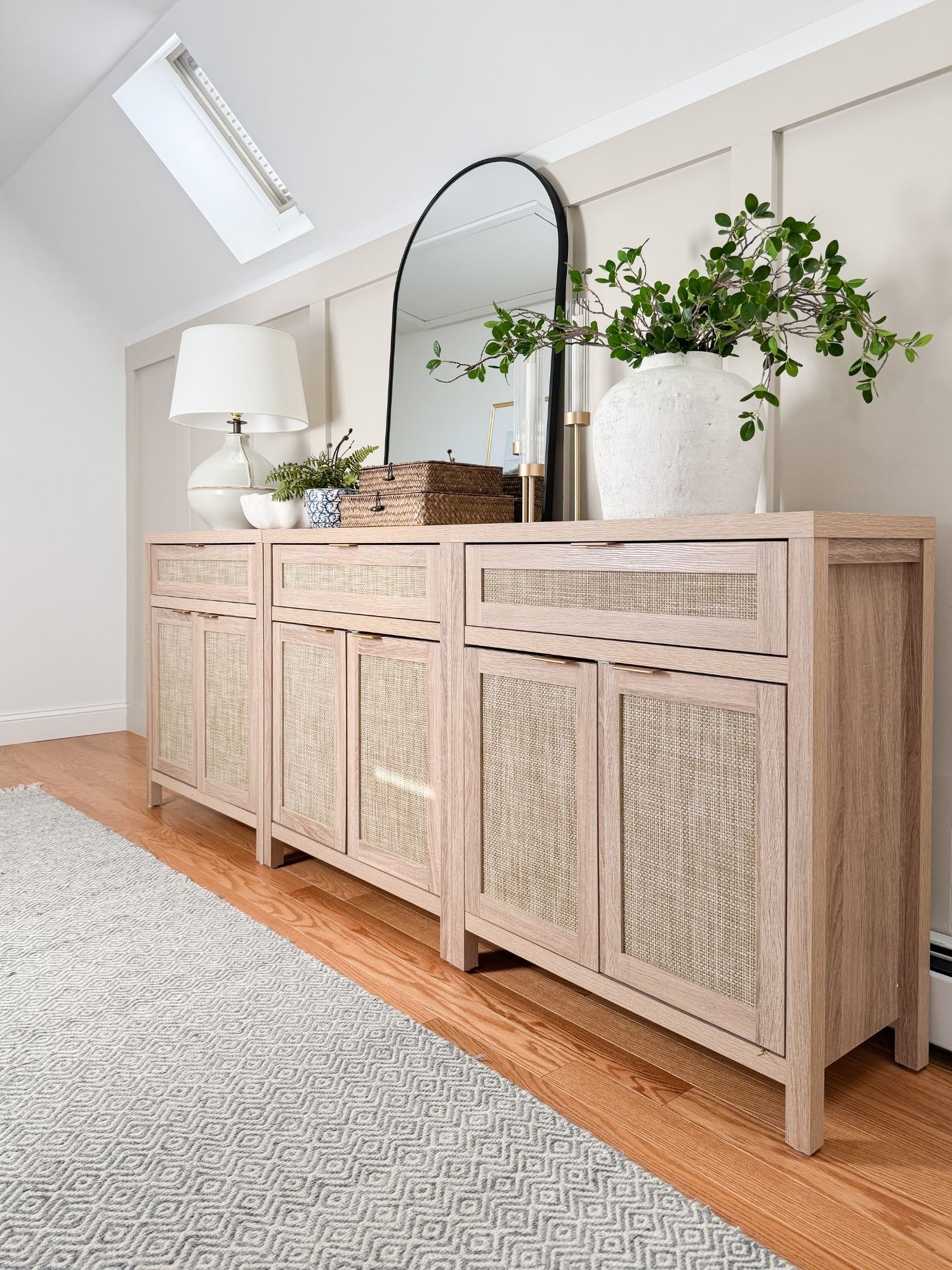 Coastal hallway vignette with light wood cabinet featuring woven cane doors, ceramic vase with greenery, arched mirror, and layered decor.
