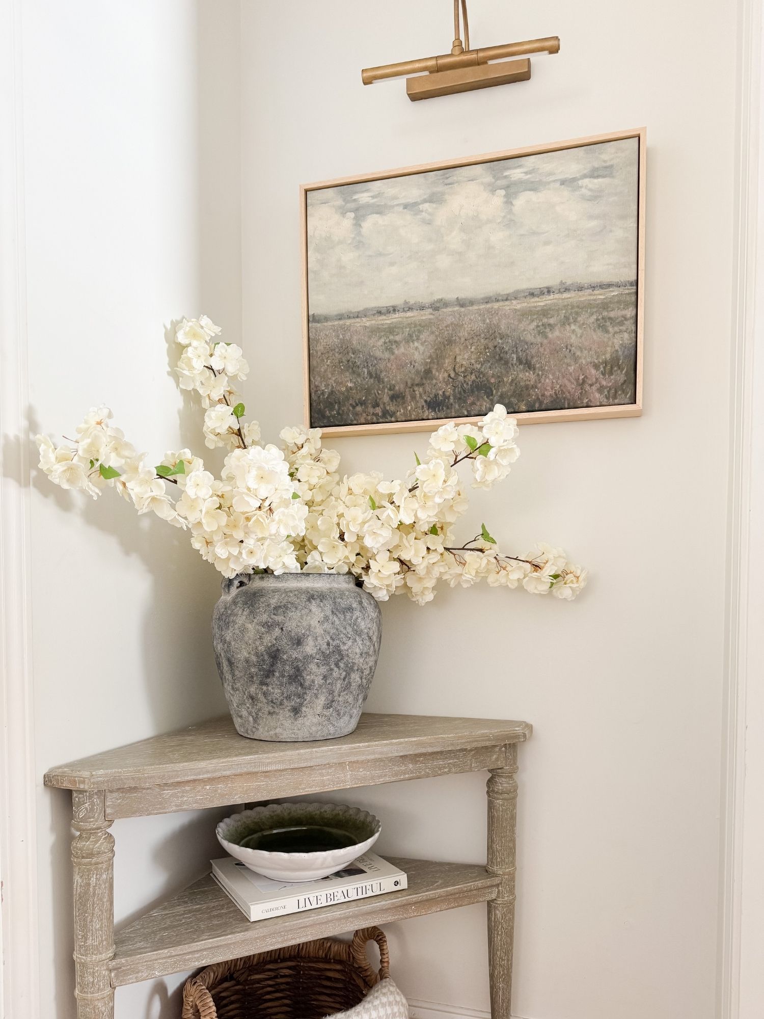 Spring hallway decor with distressed wood console table, stone vase with white florals, neutral artwork, and warm brass picture light.