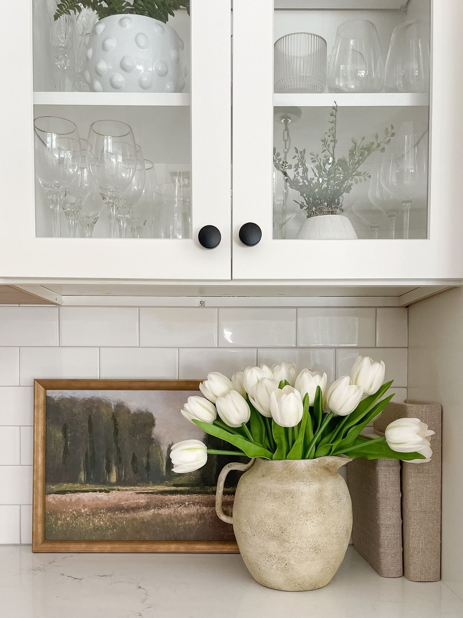 Neutral coastal kitchen decor with faux tulips in stone vase, linen decorative books, glass cabinet styling, and light coastal home accents.