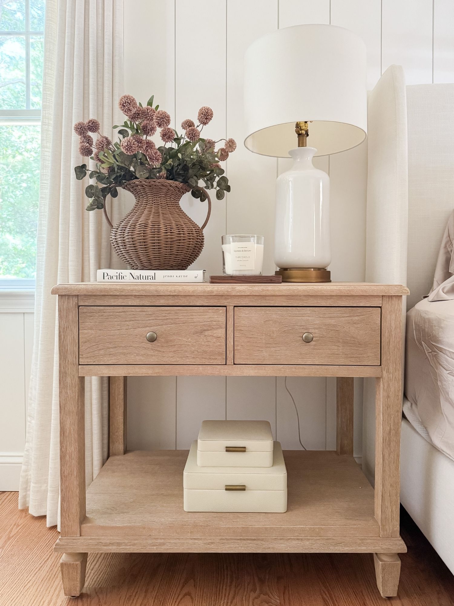 Coastal bedroom nightstand styled with woven vase, silk mums, neutral lamp, and linen decor creating a relaxed coastal bedroom aesthetic.